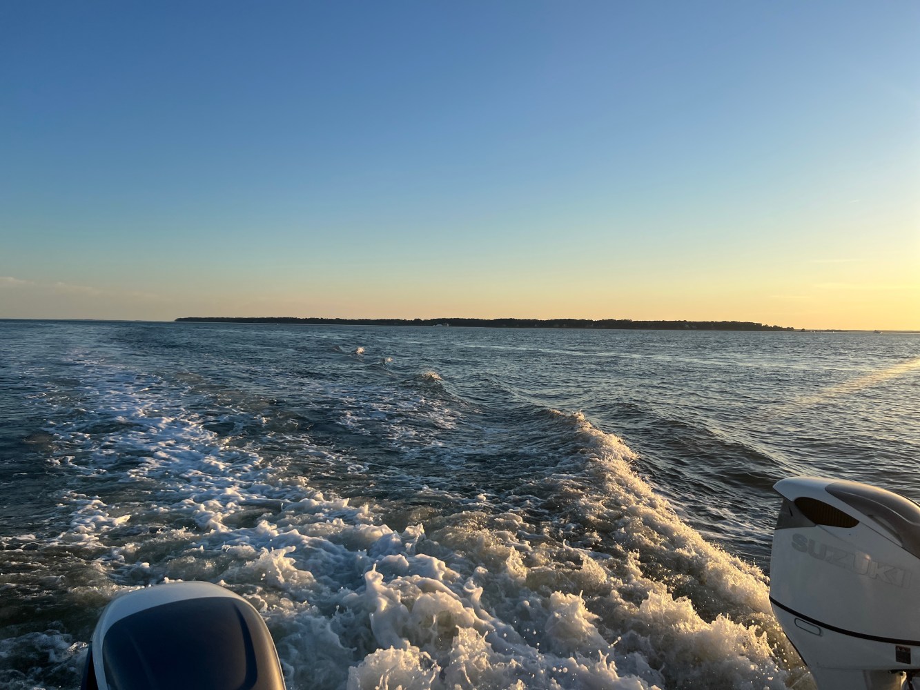 Boat wake in water with distant land under clear sky at sunset.