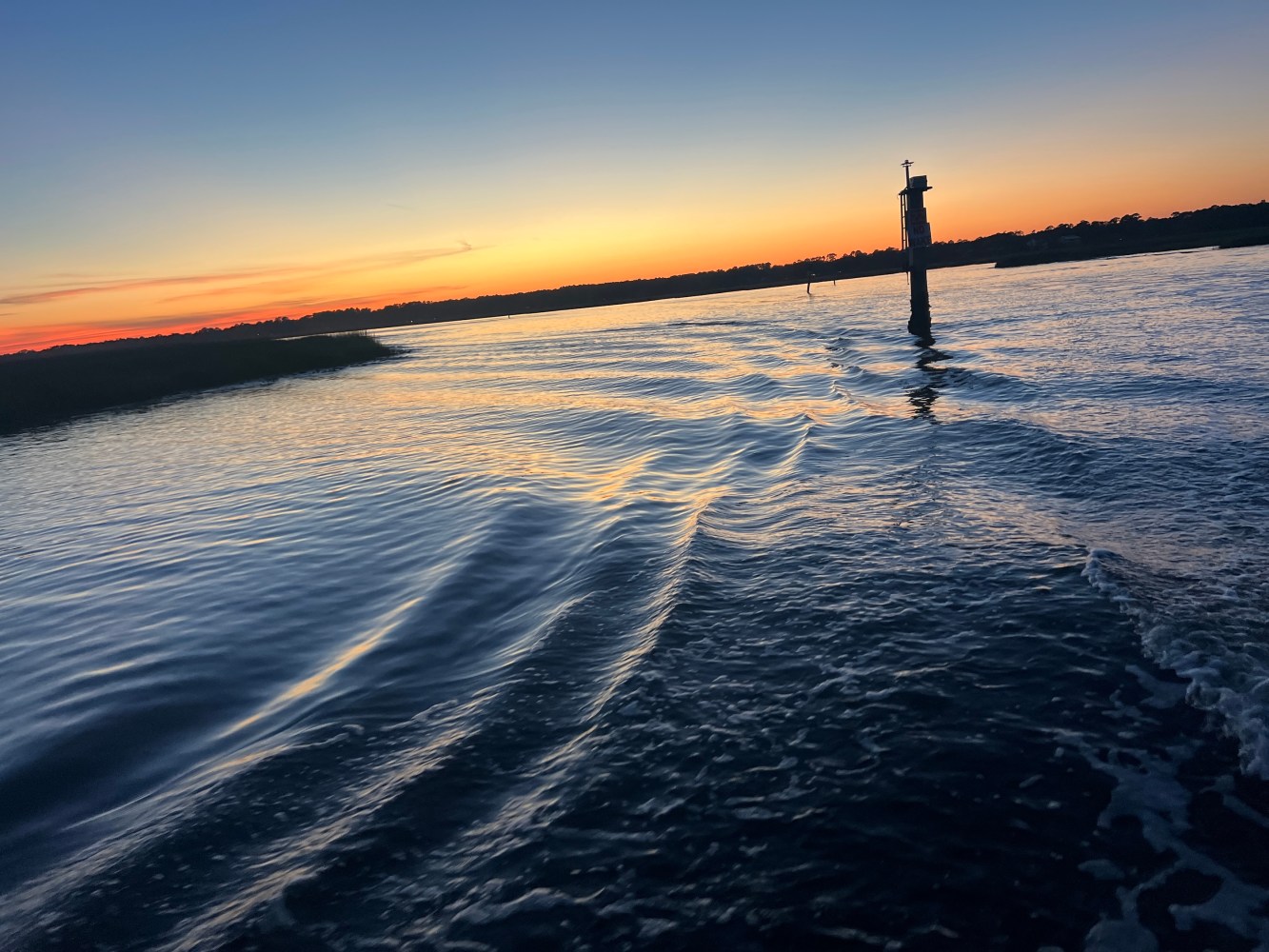 Sunset over a calm river with gentle waves and a silhouetted marker post.