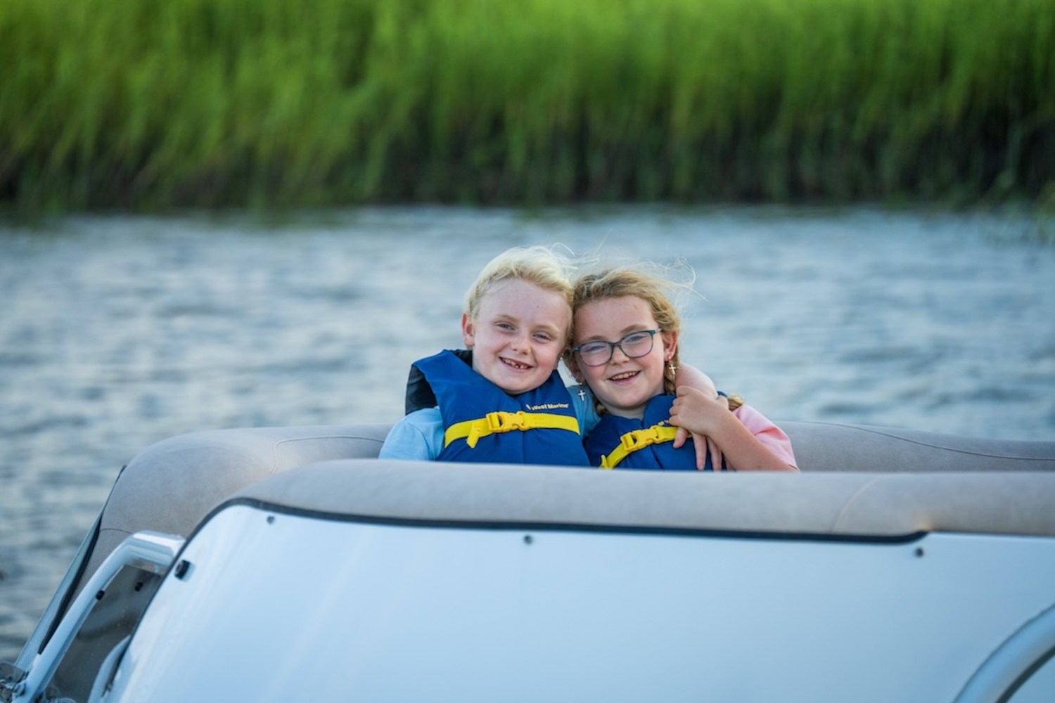 Two children in life jackets smiling on a boat with water and grass in the background.