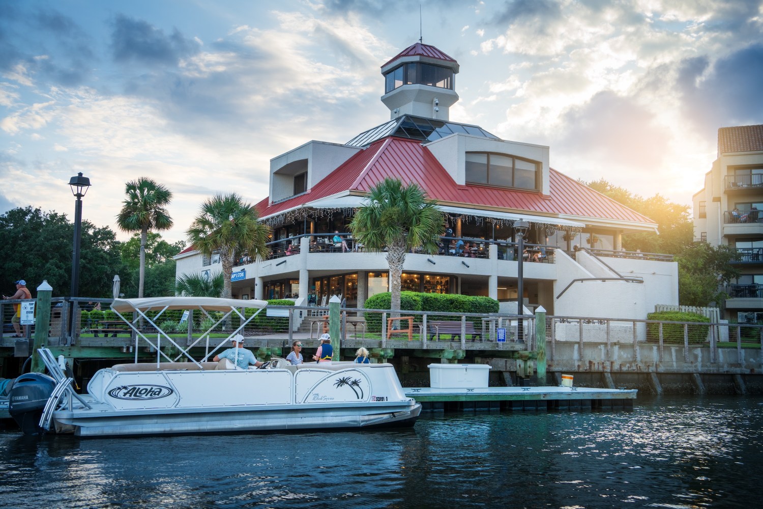 Boat docked by a waterfront restaurant with red roof at sunset.