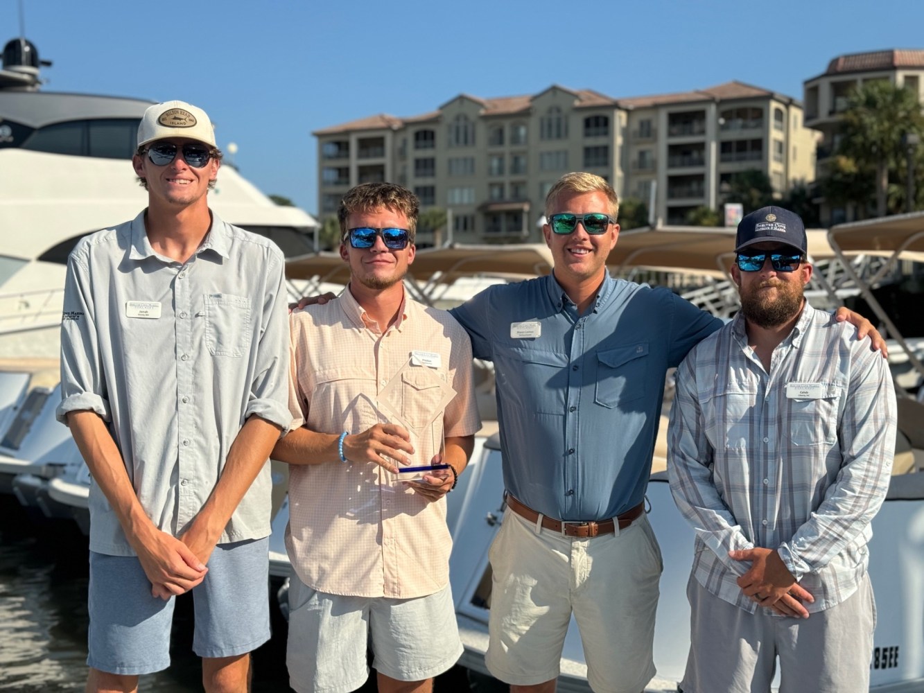 Four men in casual attire standing together in front of boats and buildings.