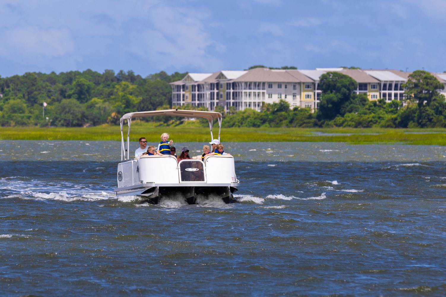 Pontoon boat on water with people, distant buildings and greenery in background.